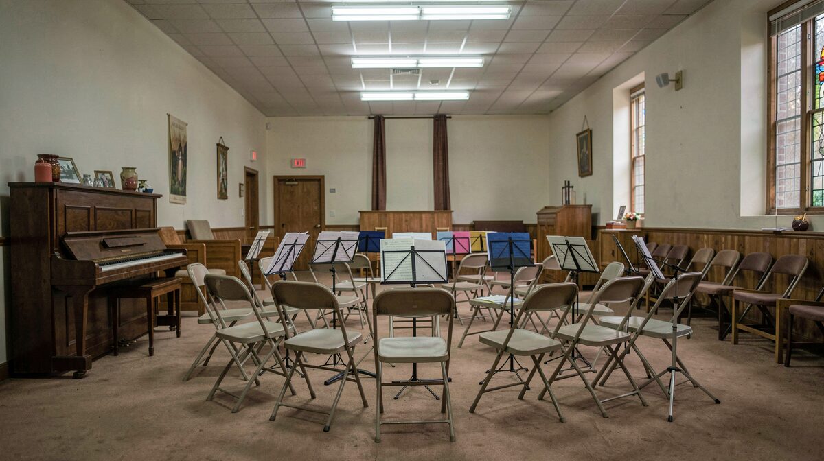 A semicircle of folding chairs and music stands in a church hall on a Tuesday night, a upright piano at the left, fluorescent light from above, wide shot from the back of the room.